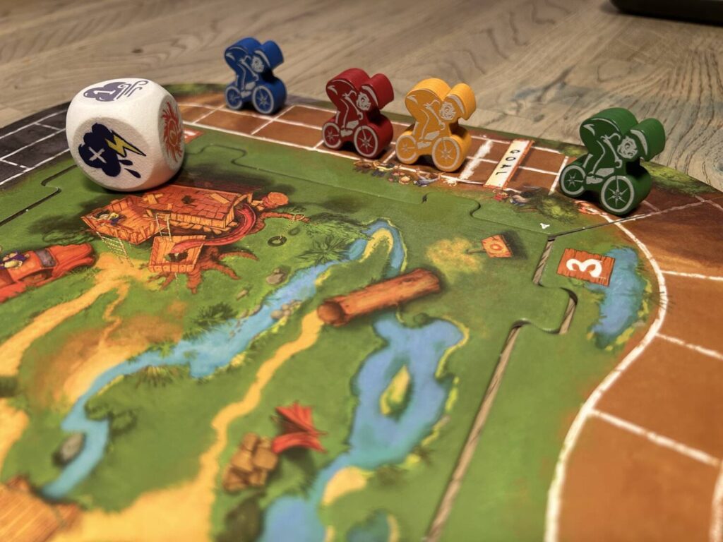 Four wooden motocross riders, each in a bright primary color, scream down the race track, the large weather die in the foreground.